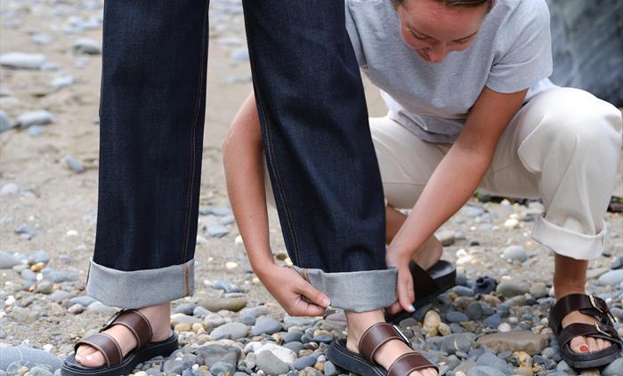 A person crouches on a rocky beach, adjusting the cuff of another’s dark raw denim jeans. Both wear sandals.  The image captures the care and styling